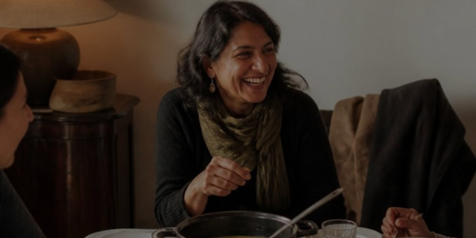 easy-entertaining-hosting-tips-hero A woman laughs at a lamp-lit table with friends, a pot of soup with a ladle in front of her.