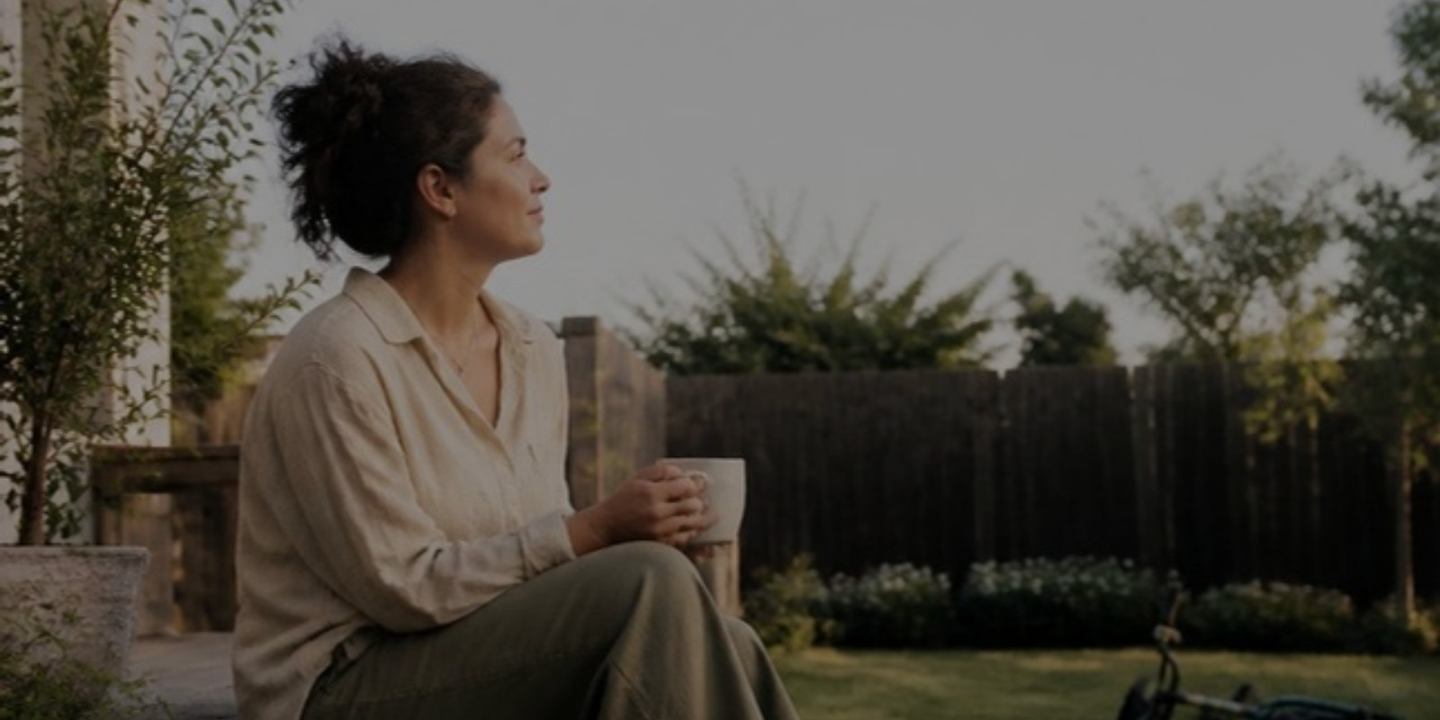 missing-life-before-kids-hero A woman sits on a garden step in soft evening light, holding a mug and looking out across the yard.