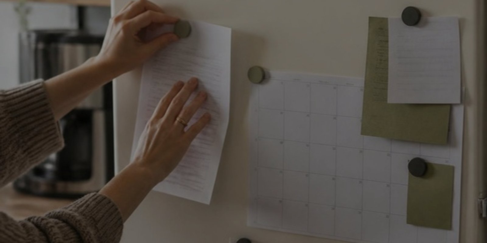 Hands pinning a note to a fridge beside a monthly calendar and lists